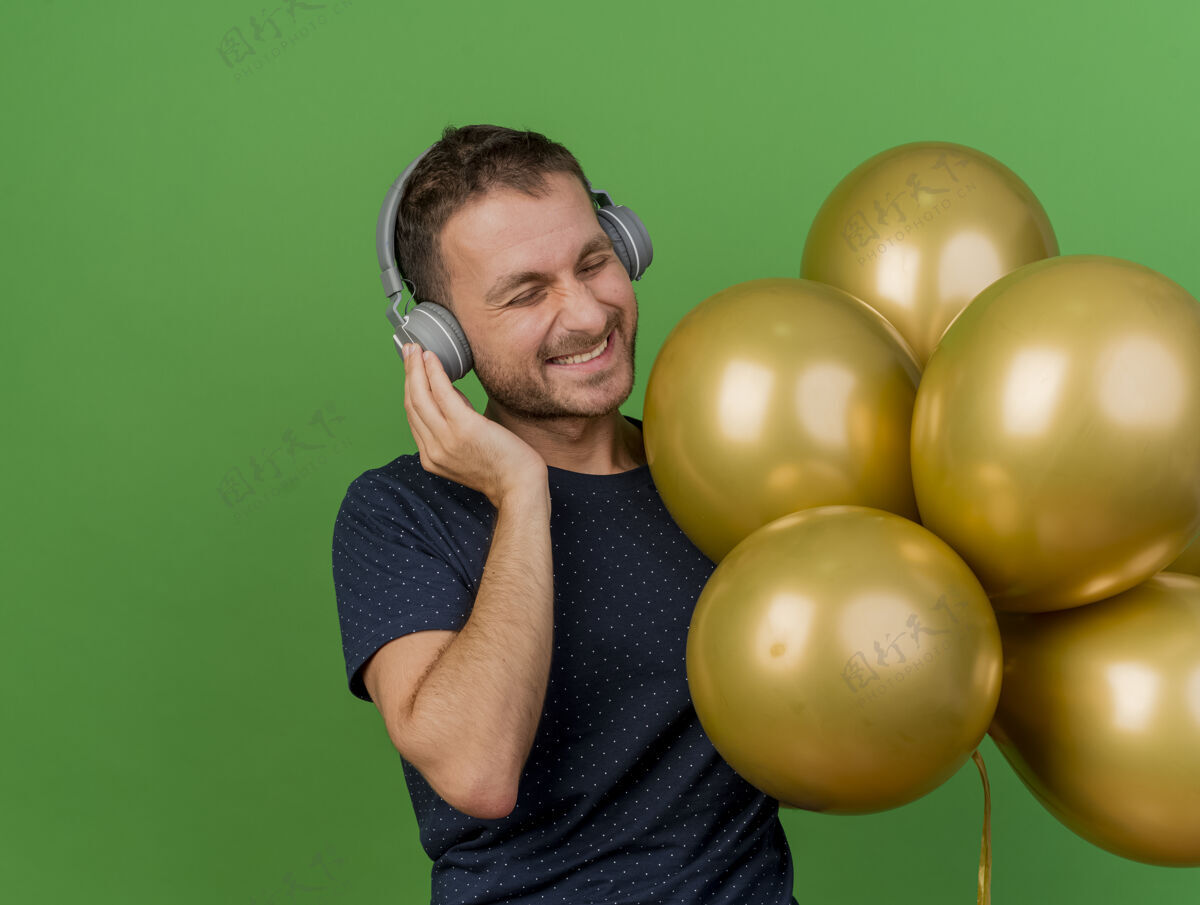 帅气戴着耳机的快乐英俊的白种人手里拿着氦气球 在绿色背景上与复制空间隔离开来氦气气球耳机