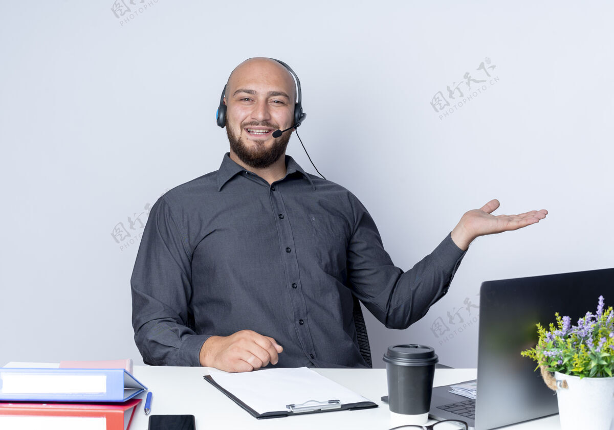 工具令人印象深刻的年轻秃头呼叫中心男子戴着耳机坐在办公桌上 工作工具显示空手孤立在白色电话中心桌子