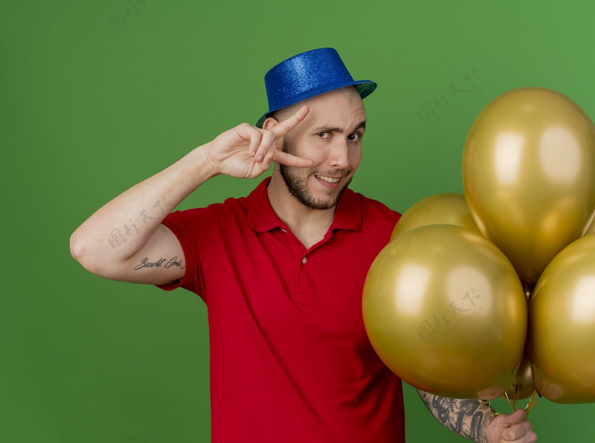 气球面带微笑的年轻英俊的斯拉夫党人戴着党的帽子拿着气球做和平标志看着隔离在绿色背景上的相机派对年轻帽子