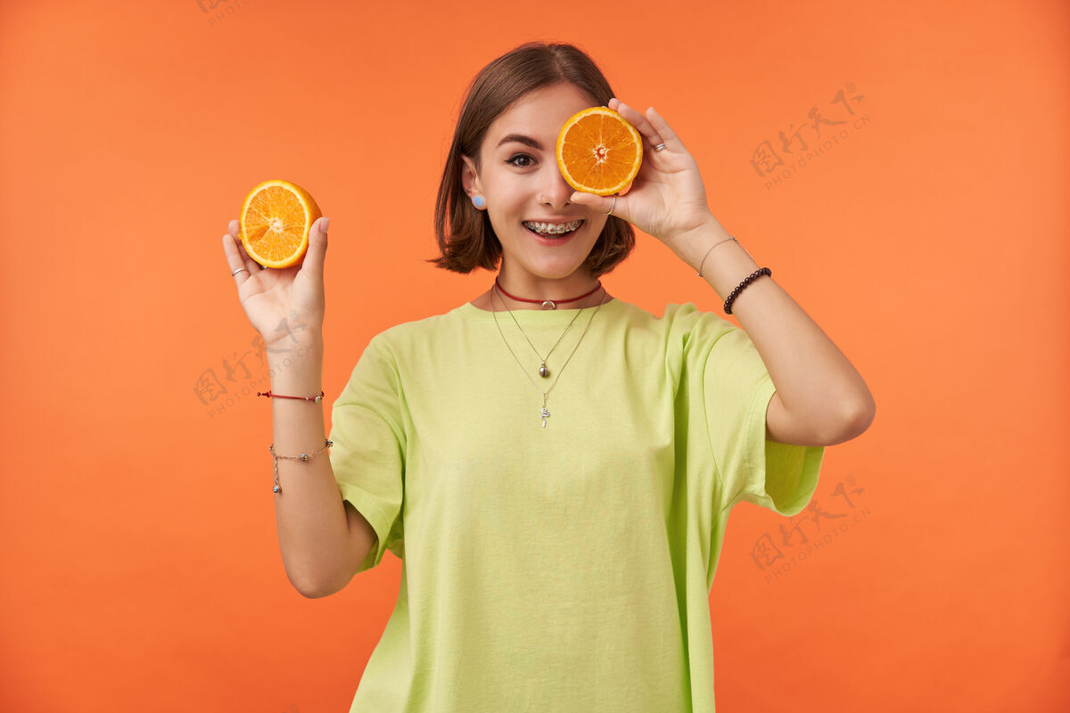 成人十几岁的小女孩 快乐快乐 深色短发遮住眼睛 遮住一只眼睛站在橙色的墙上穿着绿色t恤 戴着牙套和手镯穿洞女士热带