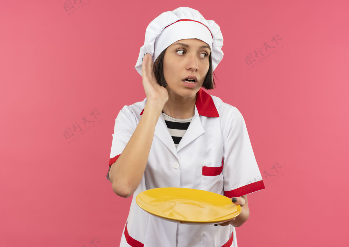 空好奇的年轻女厨师穿着厨师制服拿着空盘子看着旁边 做着听不到你的手势孤立在粉红色的复制空间女年轻拿着