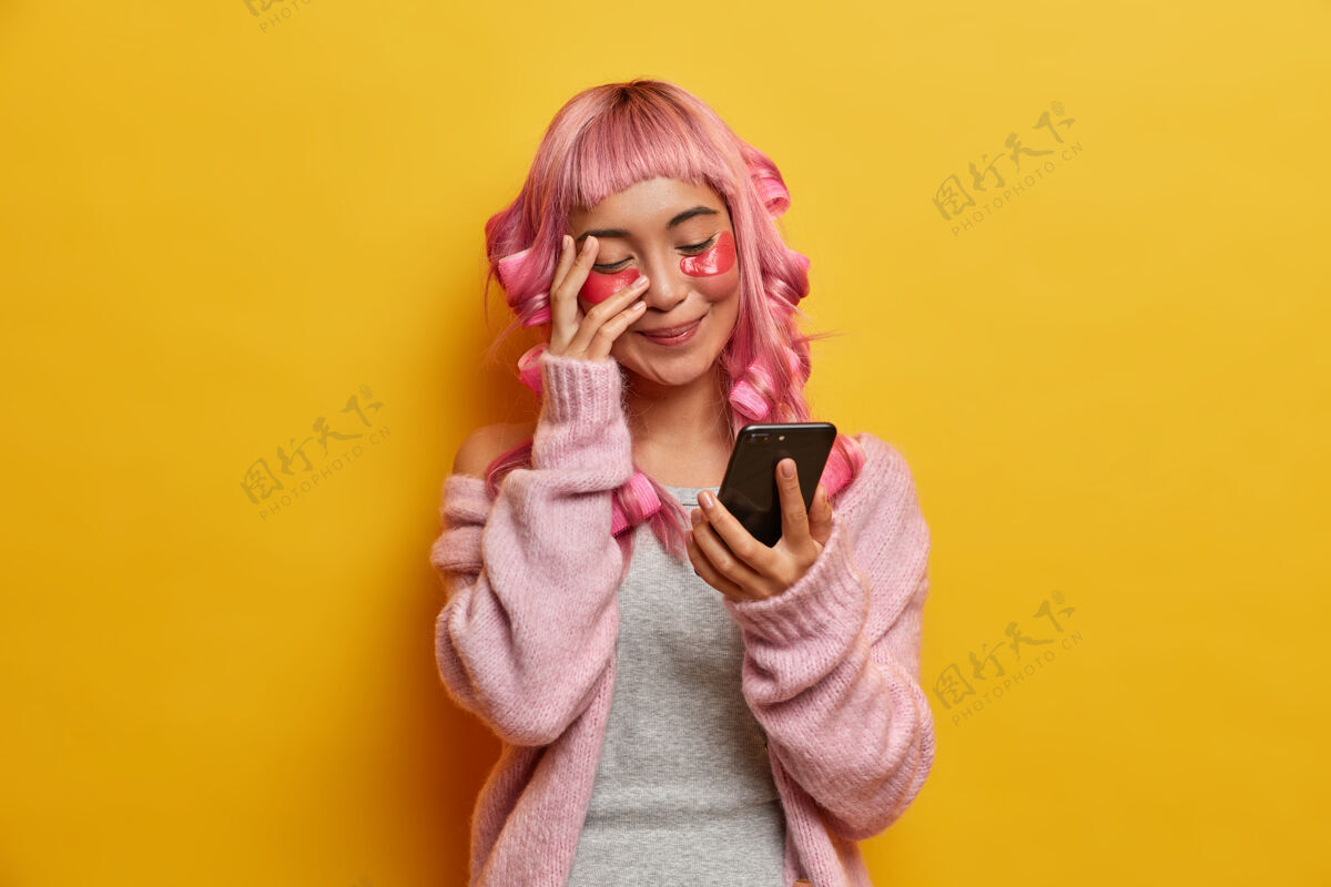 亚洲人积极的年轻女子积极地咯咯笑着 看着智能手机屏幕 读有趣的新闻 有着粉红色的长发 做发型 关心皮肤粉色人物信息