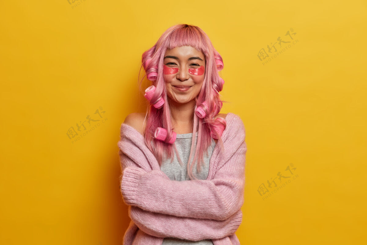美容高兴的亚洲女孩穿着羊毛套头衫感到舒适 拥抱自己 微笑温柔 有粉红色的头发 用卷发器做发型 应用美容垫染色粉色垫子