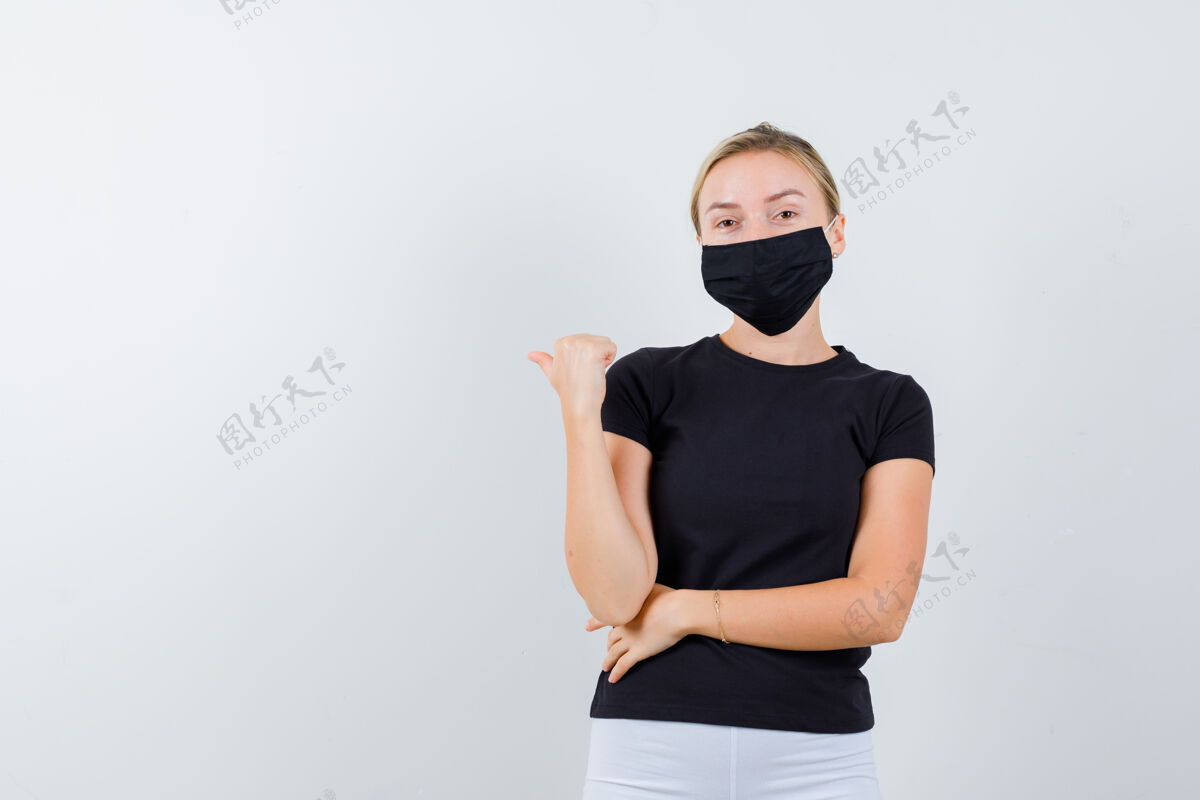 女孩一幅年轻女士的画像 她穿着黑色t恤 戴着面具 拇指向后指 看起来很自信面具模特女人