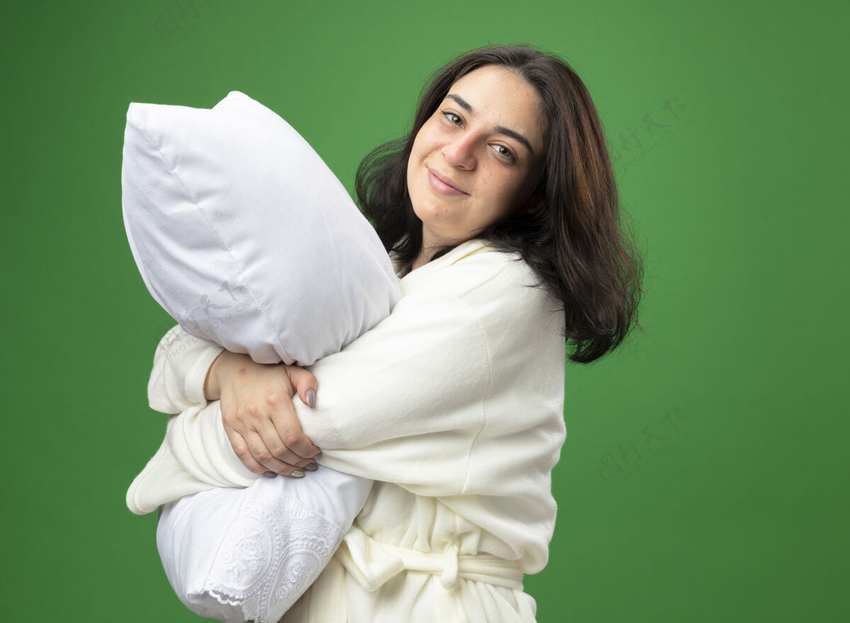 视图高兴的年轻白种人生病的女孩穿着长袍站在侧视图拥抱枕头看着相机隔离在绿色背景与复制空间复制站立穿
