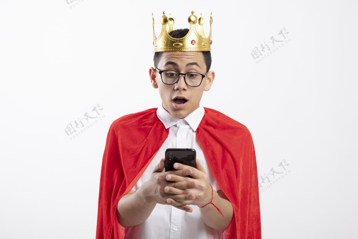 白色穿着红色斗篷 戴着眼镜 戴着皇冠的年轻超级英雄男孩惊讶地看着手机 手机被隔离在白色背景上 留着复印空间红色斗篷手机