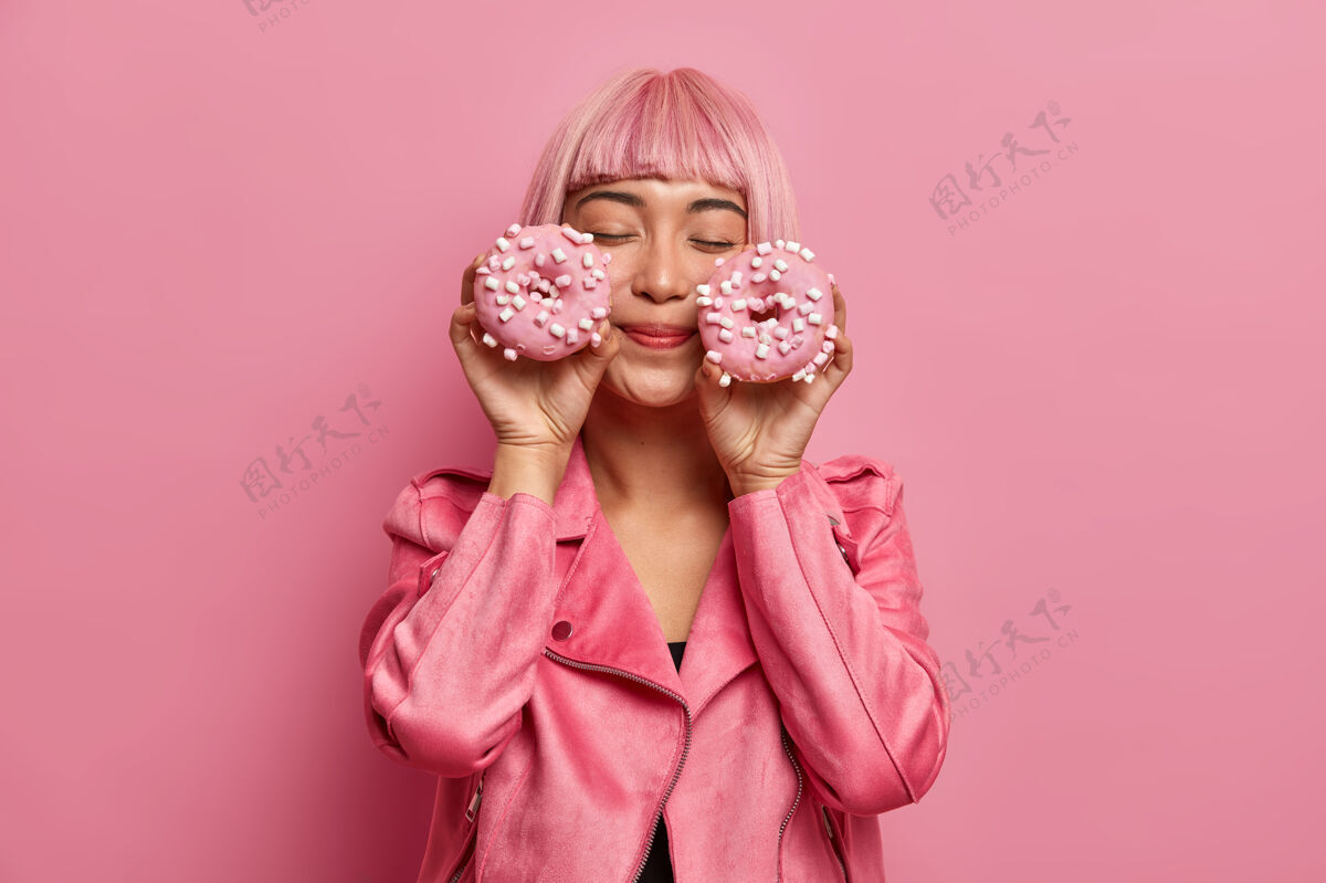 高兴满头粉红头发 留着流苏 心满意足的迷人女人 闭上眼睛 想象着甜甜圈的宜人味道 穿着玫瑰色的夹克垃圾快速糕点