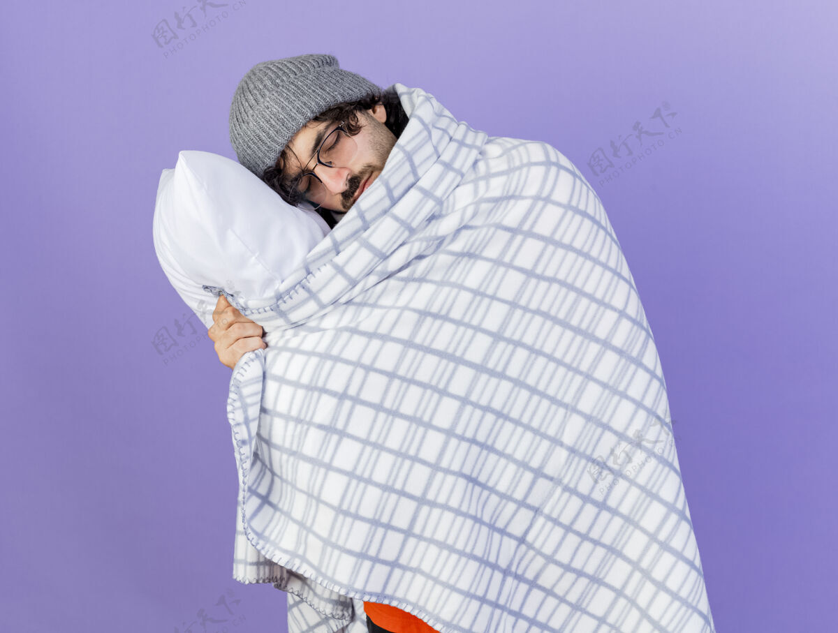 裹着昏昏欲睡的年轻白种人 戴着眼镜 戴着冬天的帽子 围着围巾 裹着格子布 抱着枕头 把头放在上面 闭着眼睛 孤立地站在紫色的背景上眼镜头闭着