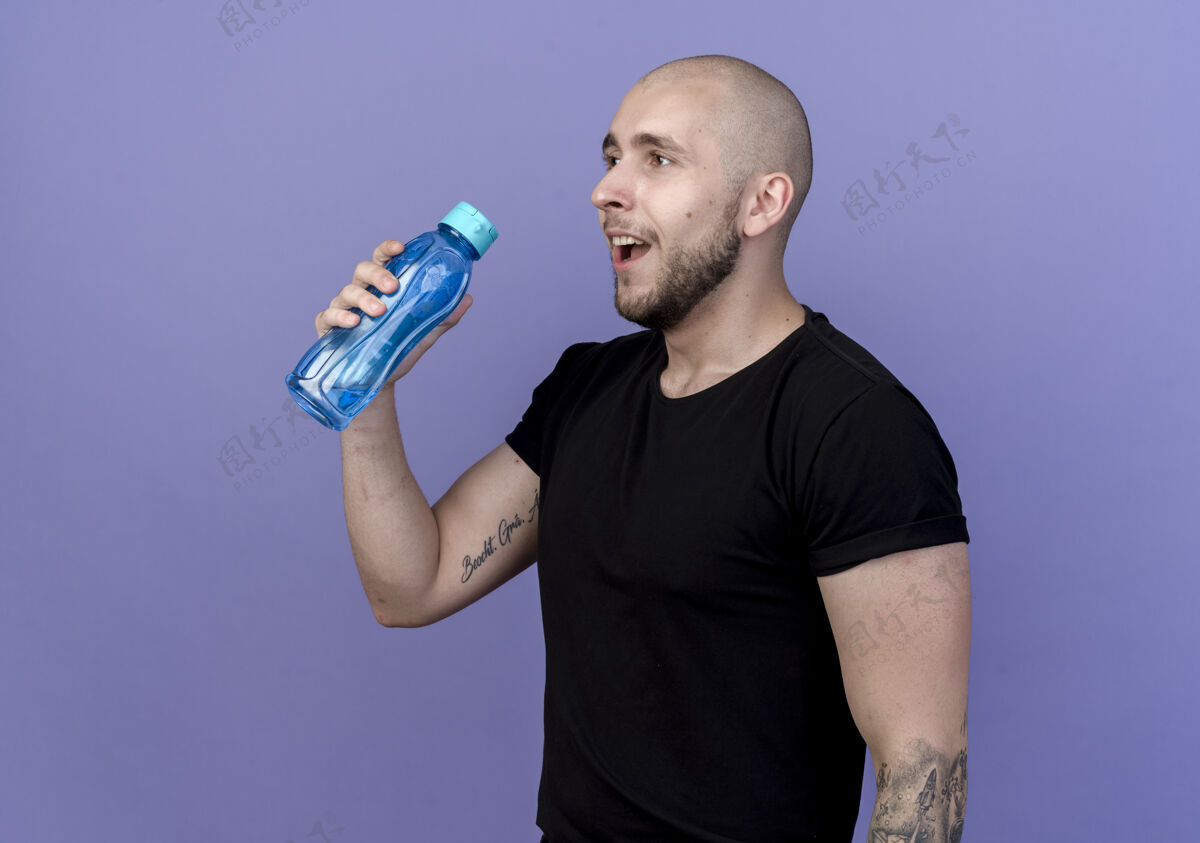 运动看着一边微笑着的年轻人 手里拿着一只孤立在紫色墙上的水瓶瓶子墙男人