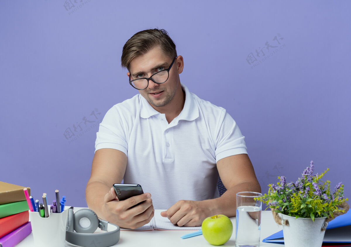 帅气年轻英俊的男学生坐在书桌旁 带着学习用具 戴着眼镜 拿着电话 孤零零地站在蓝色的墙上姿态男人穿着