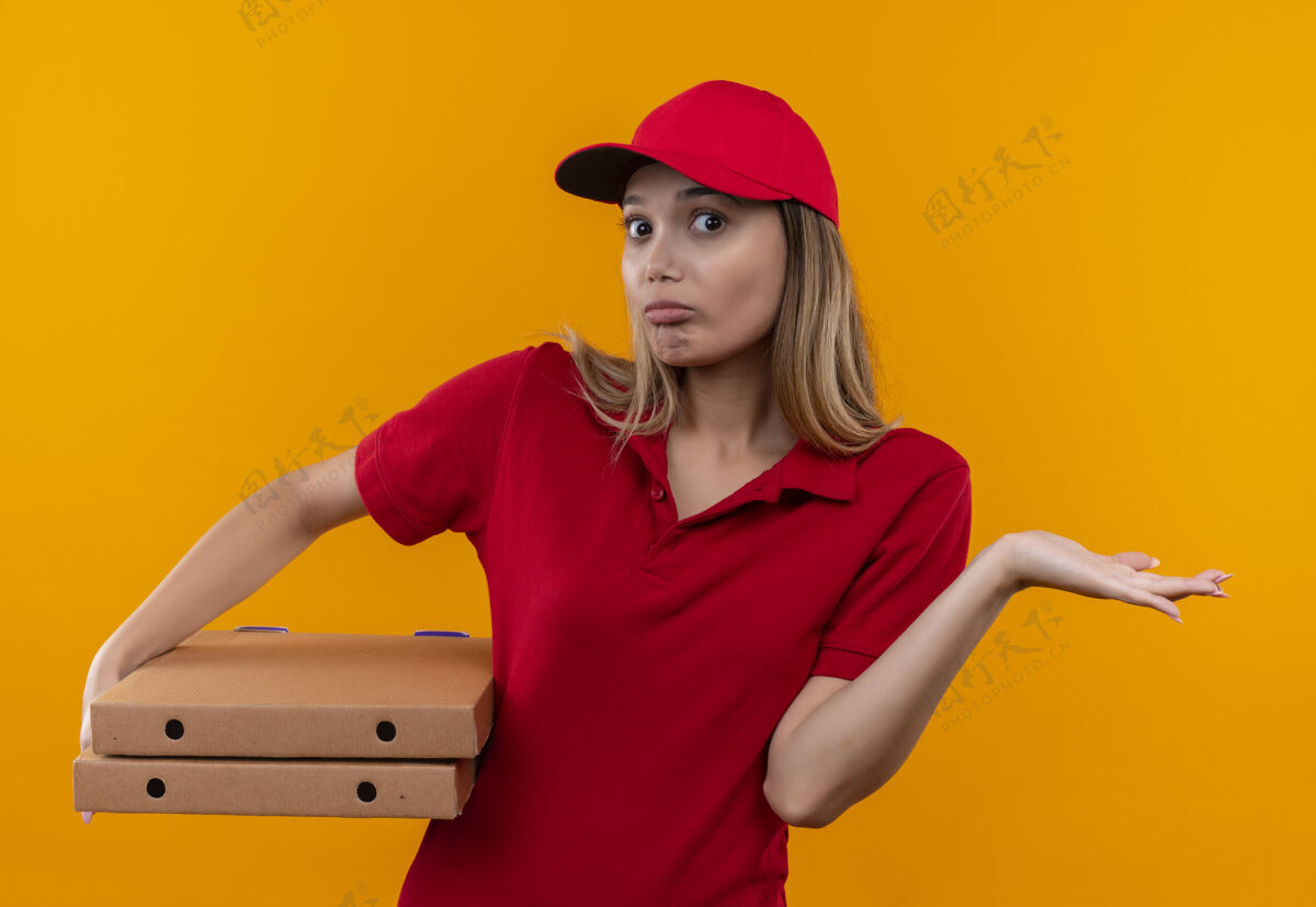 披萨困惑的年轻送货女孩穿着红色制服 戴着帽子 手里拿着披萨盒 摊开手孤立在橙色的墙上帽子送货混乱