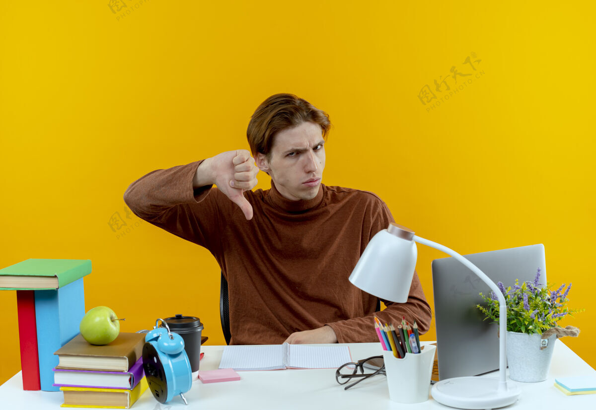 坐着一个严厉的年轻学生正坐在书桌旁 手里拿着学习用具 大拇指朝下放在黄色的桌子上年轻学校黄色