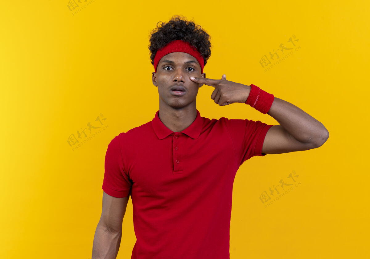 年轻年轻的美国黑人运动男子戴着头带和腕带把手指放在隔离在黄色墙上的眼睛男人头带手指