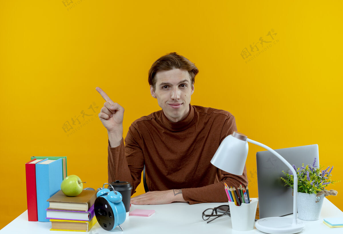 男孩高兴的小男生坐在书桌旁 用学习工具指着旁边的黄色黄色学校坐着