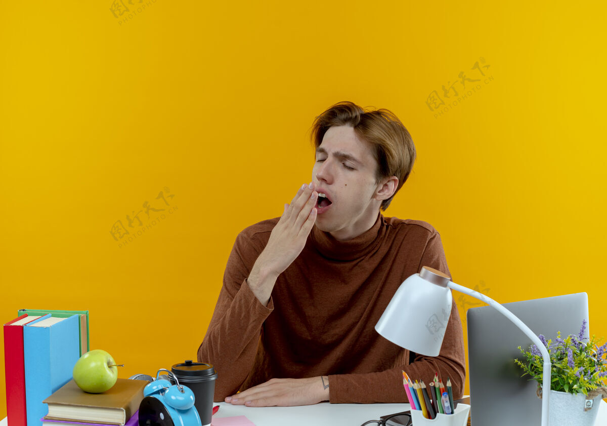 工具小男孩闭着眼睛打呵欠坐在书桌旁 手里拿着学习工具 用手捂着黄色的嘴巴手学校桌子
