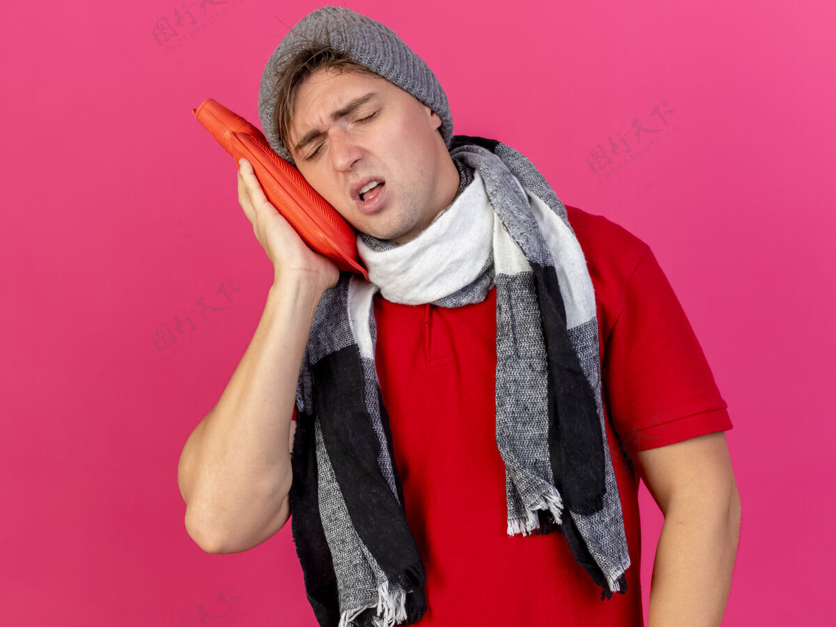 人愁眉不展的年轻英俊的金发病男子戴着冬天的帽子和围巾用热水瓶抚摸着脸闭着眼睛隔离在粉红色的墙上站眼睛触摸