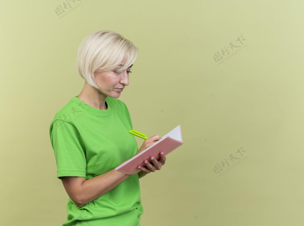 表情专注的中年金发女人站在侧视图上用橄榄绿的墙上孤立的笔在记事本上写字个人资料市民橄榄