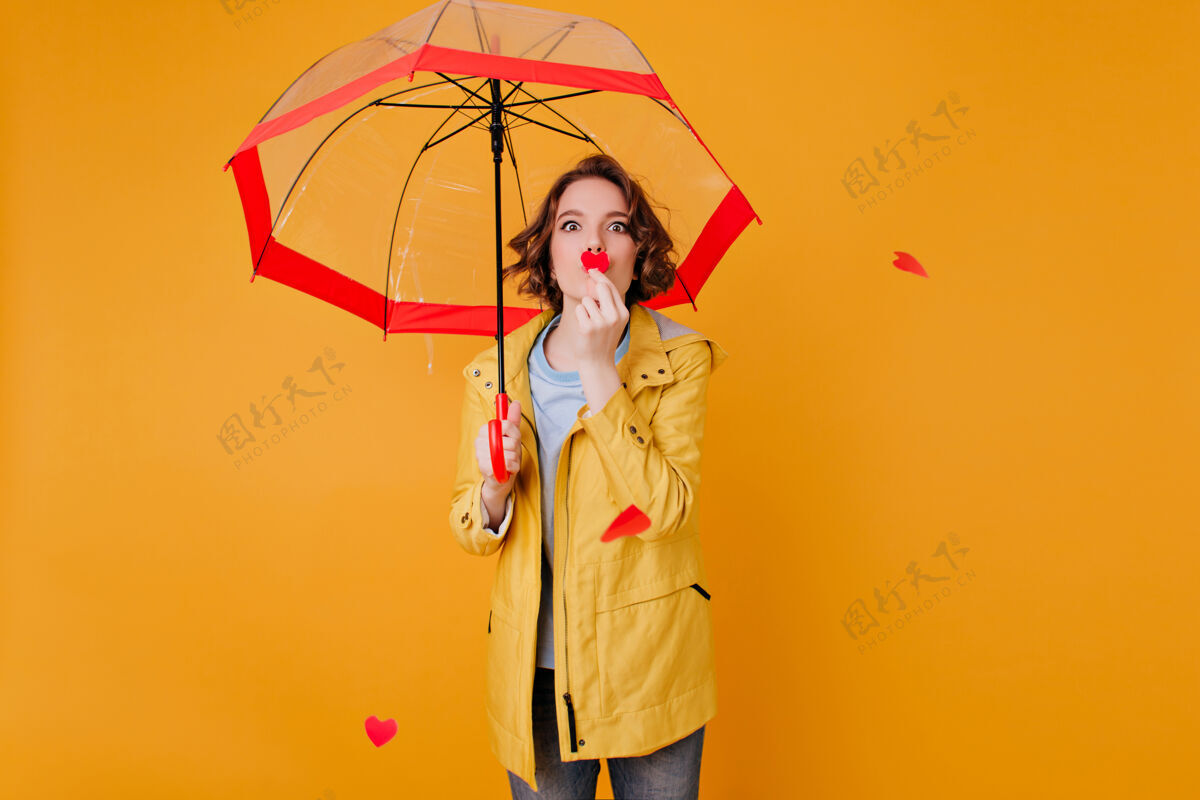 肖像可爱的白色女模特抱着小纸心在阳伞下摆姿势室内照片中 穿着黄色夹克的无忧无虑的女孩在打着伞拍照时放松优雅阳伞卷发