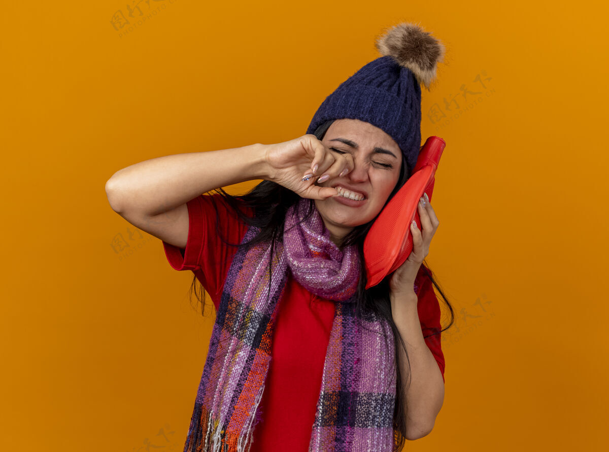 站立带着冬天帽子和围巾的疼痛的年轻生病的女人用热水袋触摸脸用闭着的眼睛触摸鼻子隔离在橙色的墙上帽子包姿势