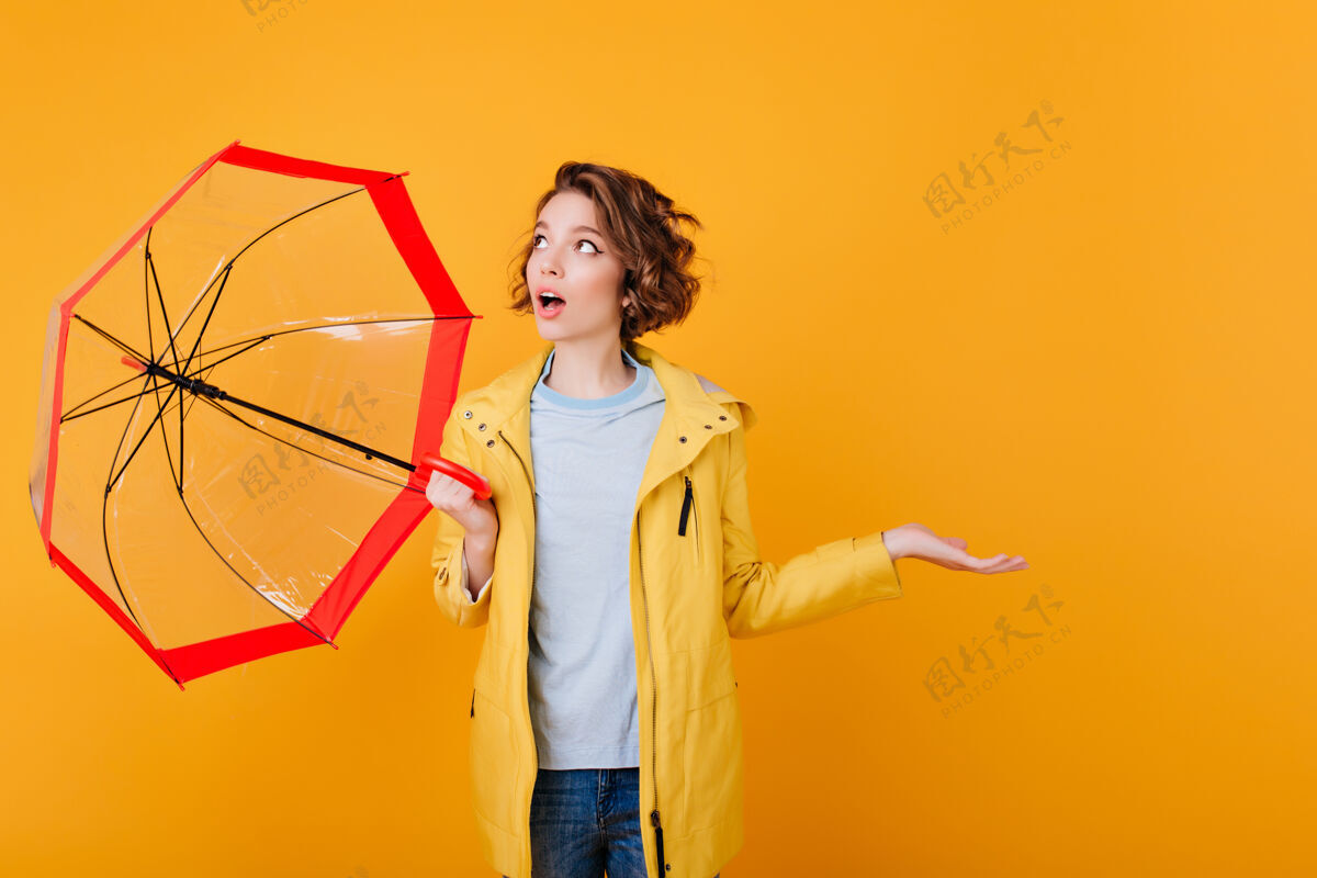 年轻穿着外套的女孩惊奇地抬起头来 手里拿着伞震惊的年轻女士用阳伞隔离在明亮的橙色墙上雨黑发室内