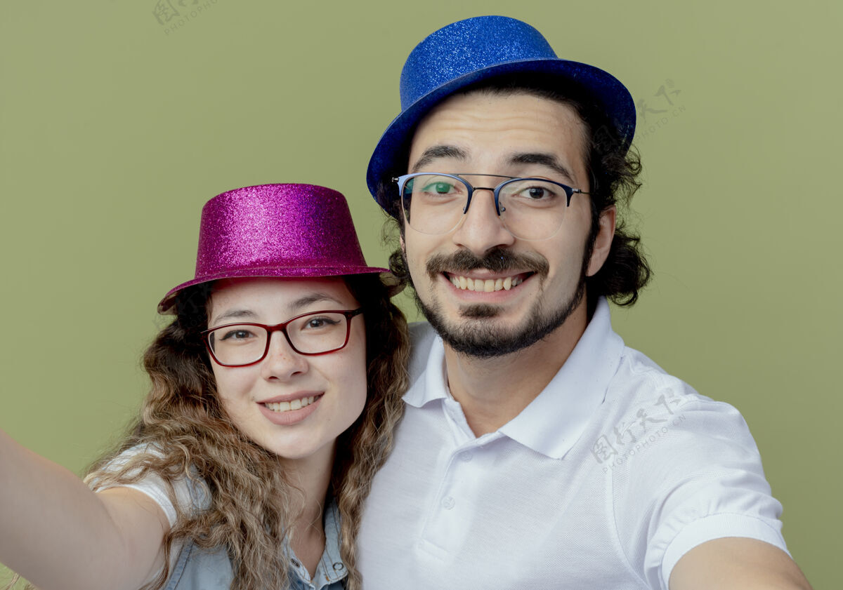 相机一对微笑的年轻夫妇戴着粉色和蓝色的帽子 拿着相机在橄榄绿上隔离情侣橄榄年轻