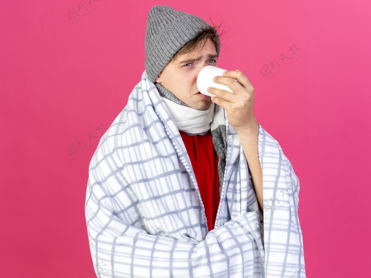 英俊皱眉的年轻英俊的金发病男子戴着冬天的帽子和围巾裹在格子布看起来直喝一杯茶隔离在粉红色的墙上市民男人格子