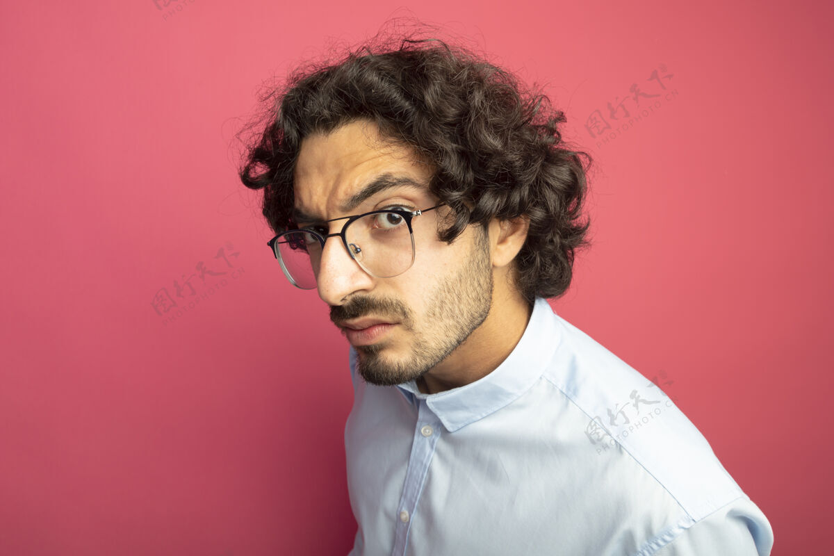 穿着特写镜头皱眉的年轻帅哥戴着眼镜站在侧面看前方隔离在粉红色的墙上立场脸感情