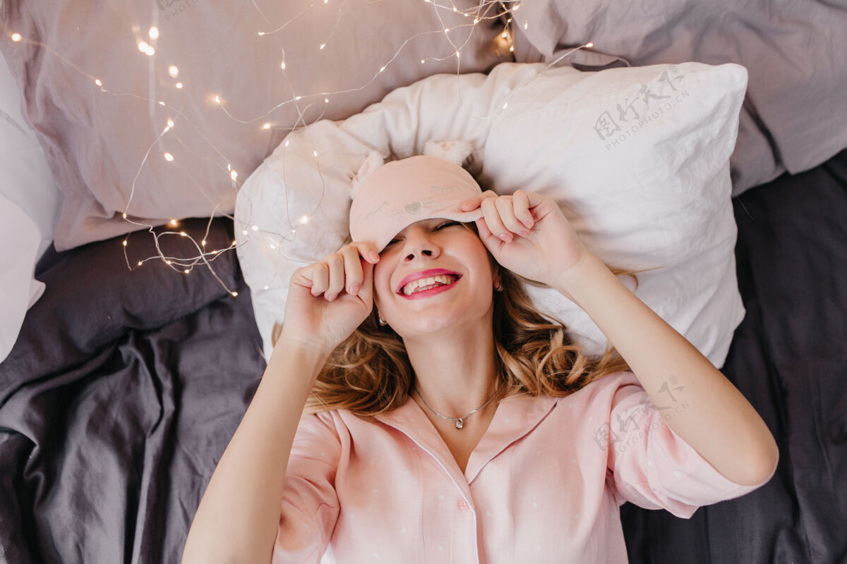 睡眠面罩可爱的穿着粉色睡衣的年轻女子微笑着在床上摆姿势戴着眼罩的高加索女孩兴奋地在早晨放松和大笑积极唤醒年轻