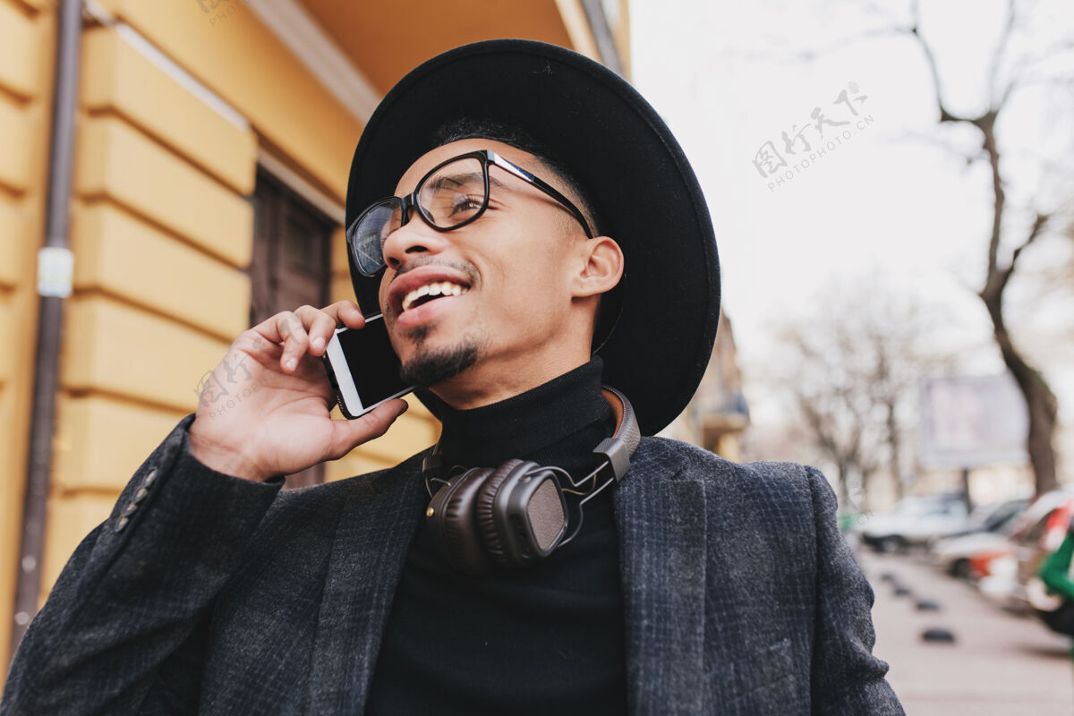 电话一个无忧无虑的非洲男人 留着短发 微笑着打电话一张热情的戴着帽子的黑人年轻人拿着手机走在街上的户外照片手机帅气忙碌
