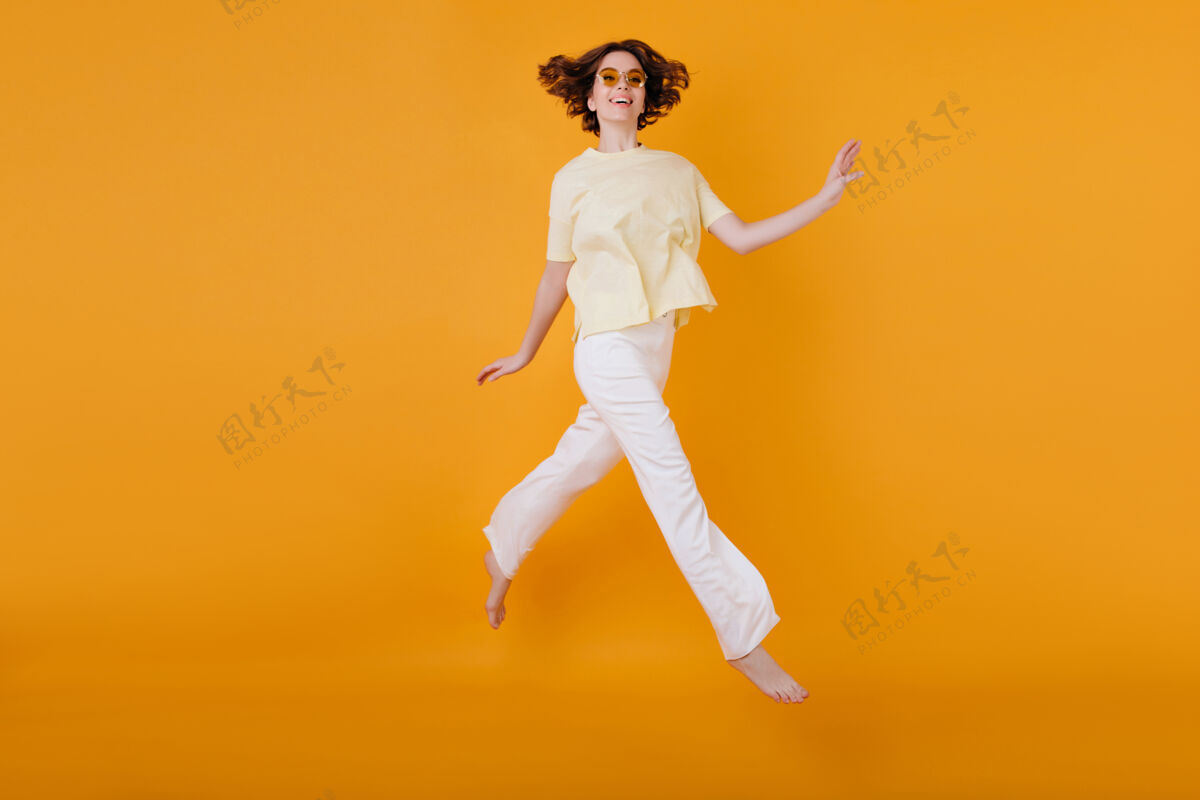 兴奋身着黄色t恤和白色裤子的快乐女孩在橙色墙上奔跑的全长肖像美丽的白种女人愉快地跳舞成人肖像情绪