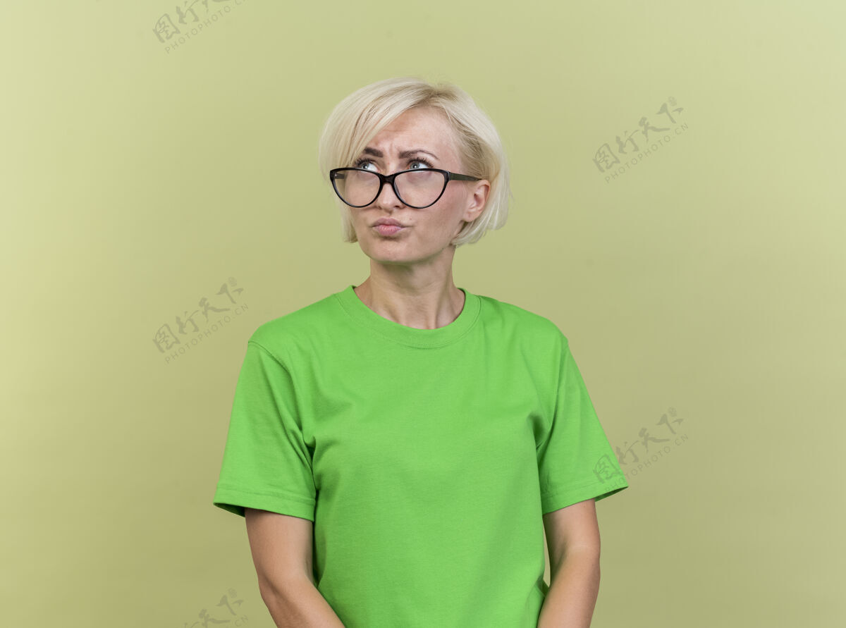 脸困惑的中年金发女人戴着眼镜 看着隔离在橄榄绿墙上的一面衣服人感觉