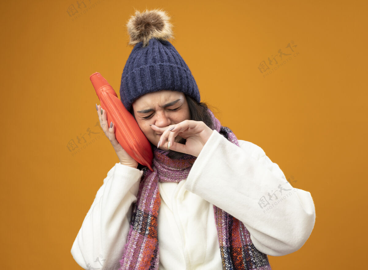 衣服愁眉苦脸的年轻病妇 戴着长袍 冬帽 围巾 用热水袋摸着头 闭着眼睛擦着鼻子 隔离在橘色的墙上头脸人