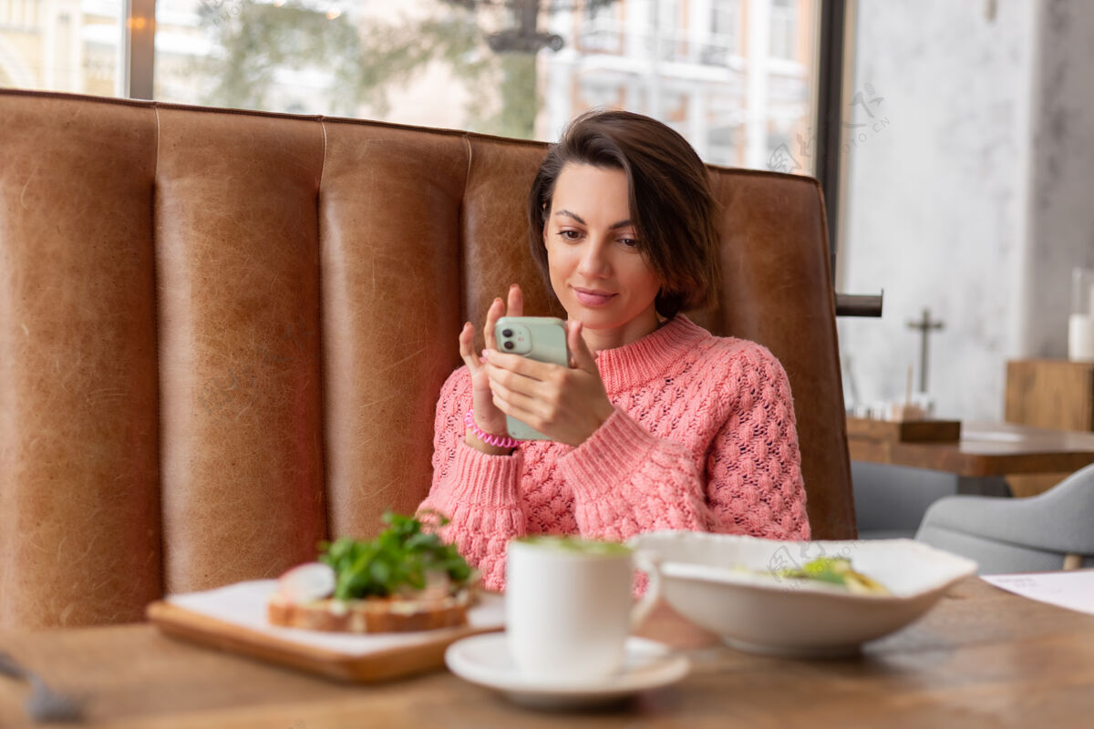 乡村餐厅里一位穿着暖和毛衣的女士正在看电话里的东西 桌上摆着美味健康的早餐和抹茶拿铁早午餐健康女孩