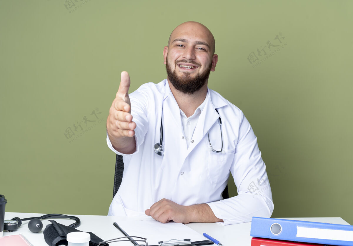 相机面带微笑的年轻秃头男医生穿着医用长袍和听诊器坐在办公桌前工作坐着医生工作
