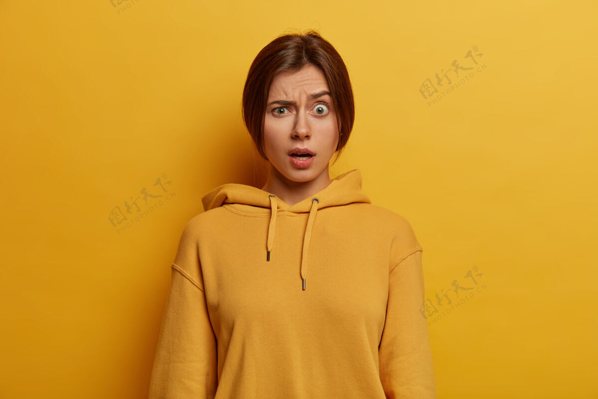 模特愤怒的欧洲年轻女子照片扬眉吐气 有意想不到的表情 笑脸 穿休闲连帽衫 表达惊奇 对着黄墙摆姿势人脸表情概念沮丧困惑休闲