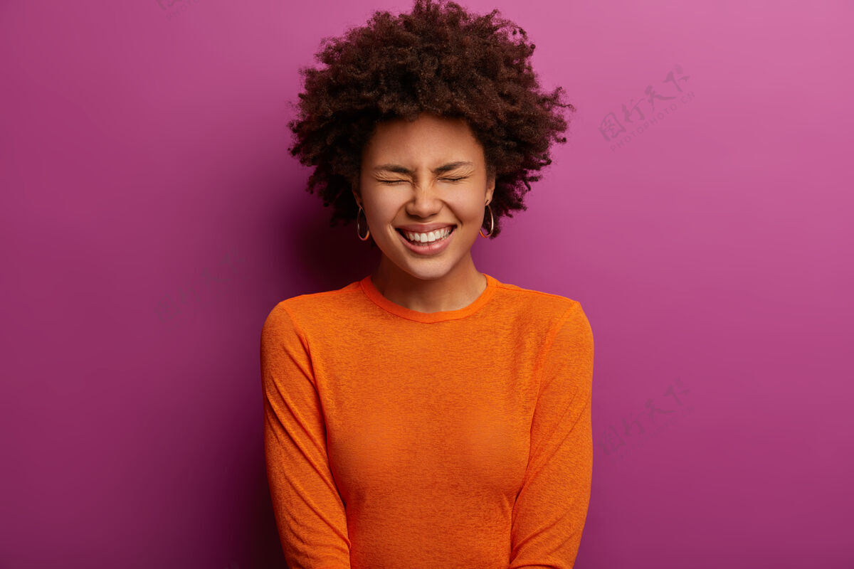 年轻民族喜出望外的年轻女子笑着积极的东西 闭上眼睛 高兴地笑着 穿着橙色套头衫 听到有趣的笑话 对充满活力的紫色墙摆姿势情感概念表情微笑乐观