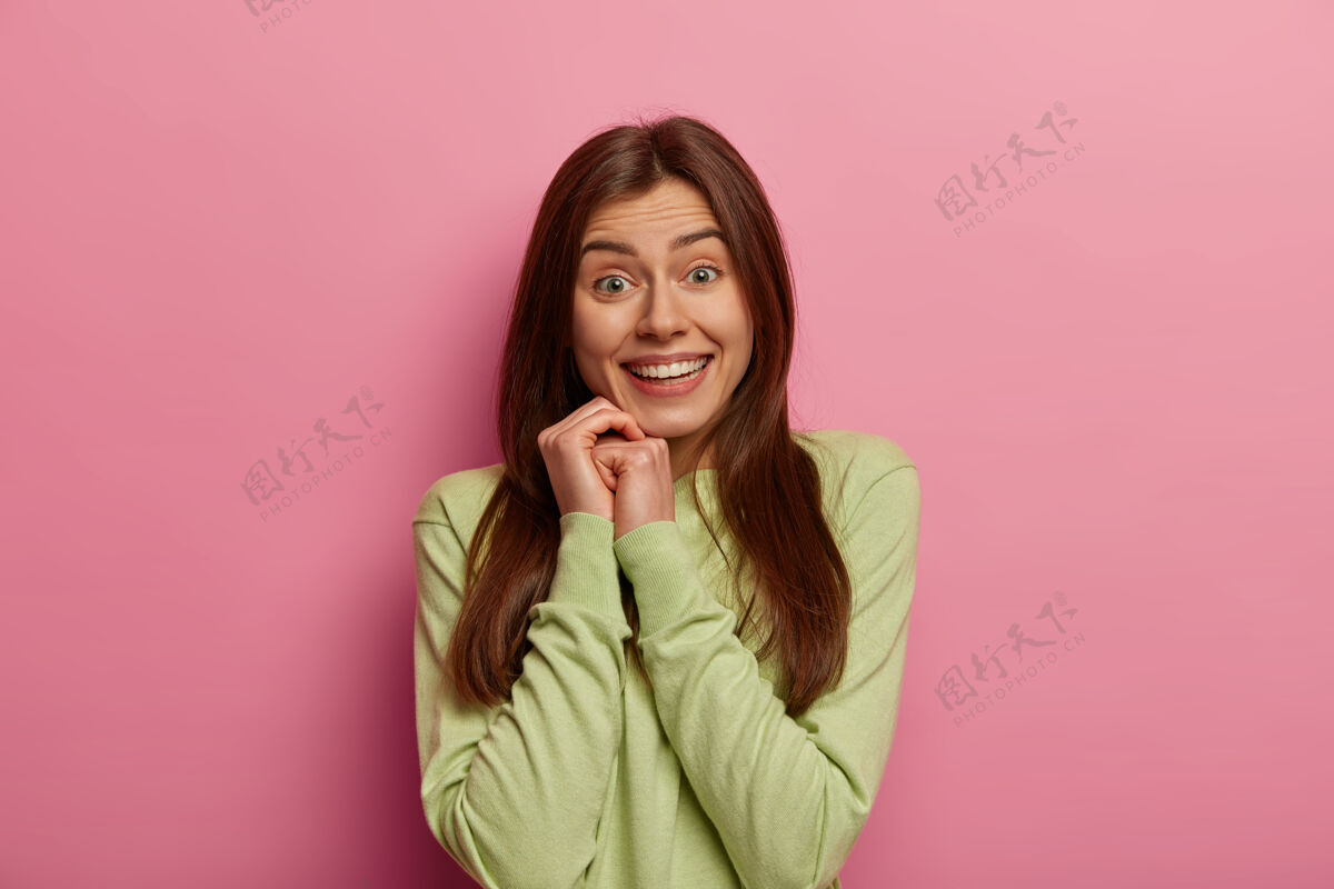 积极室内拍摄的漂亮黑发欧洲女人双手贴脸 微笑温柔 拥有洁白完美的牙齿 健康的皮肤 穿着绿色套头衫 对着粉彩玫瑰色的墙壁摆姿势幸福欧洲女人温柔