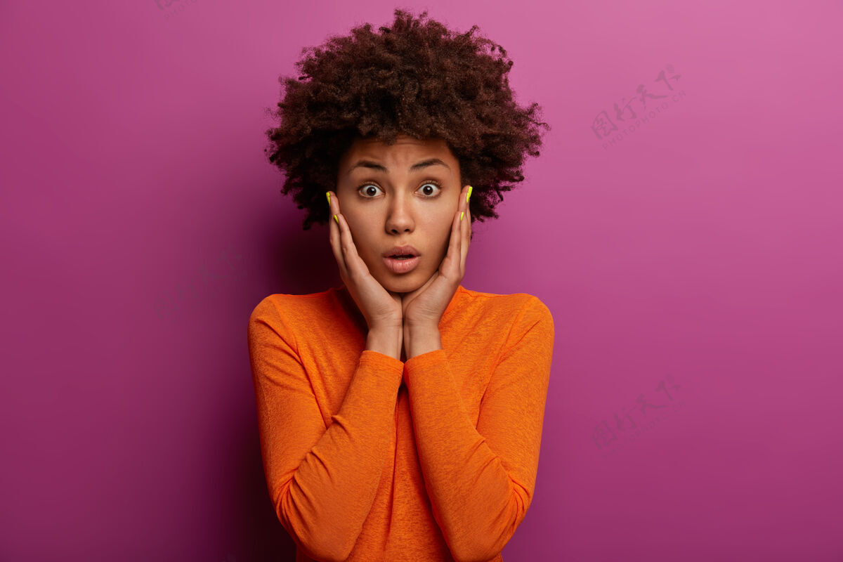 美国迷人的非裔美国妇女抓住脸颊 穿着橙色套头衫 意识到她忘记了一些重要的事情 收到大罚款 对紫色的墙壁摆姿势人类的面部表情恐惧惊讶卷发