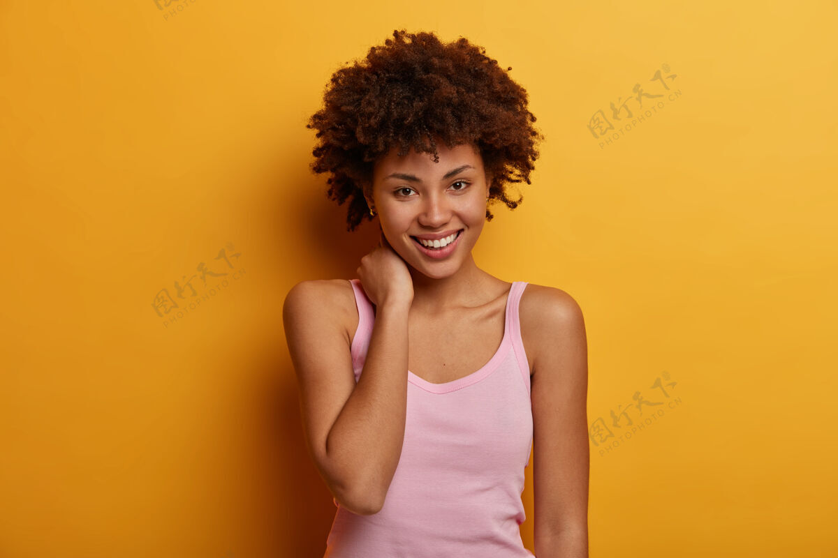 微笑可爱的年轻女子抚摸着脖子 面带温柔的微笑 感觉良好和感动 穿着休闲t恤 目光直视 隔离在黄色的墙壁上积极的人脸表情魅力满足人