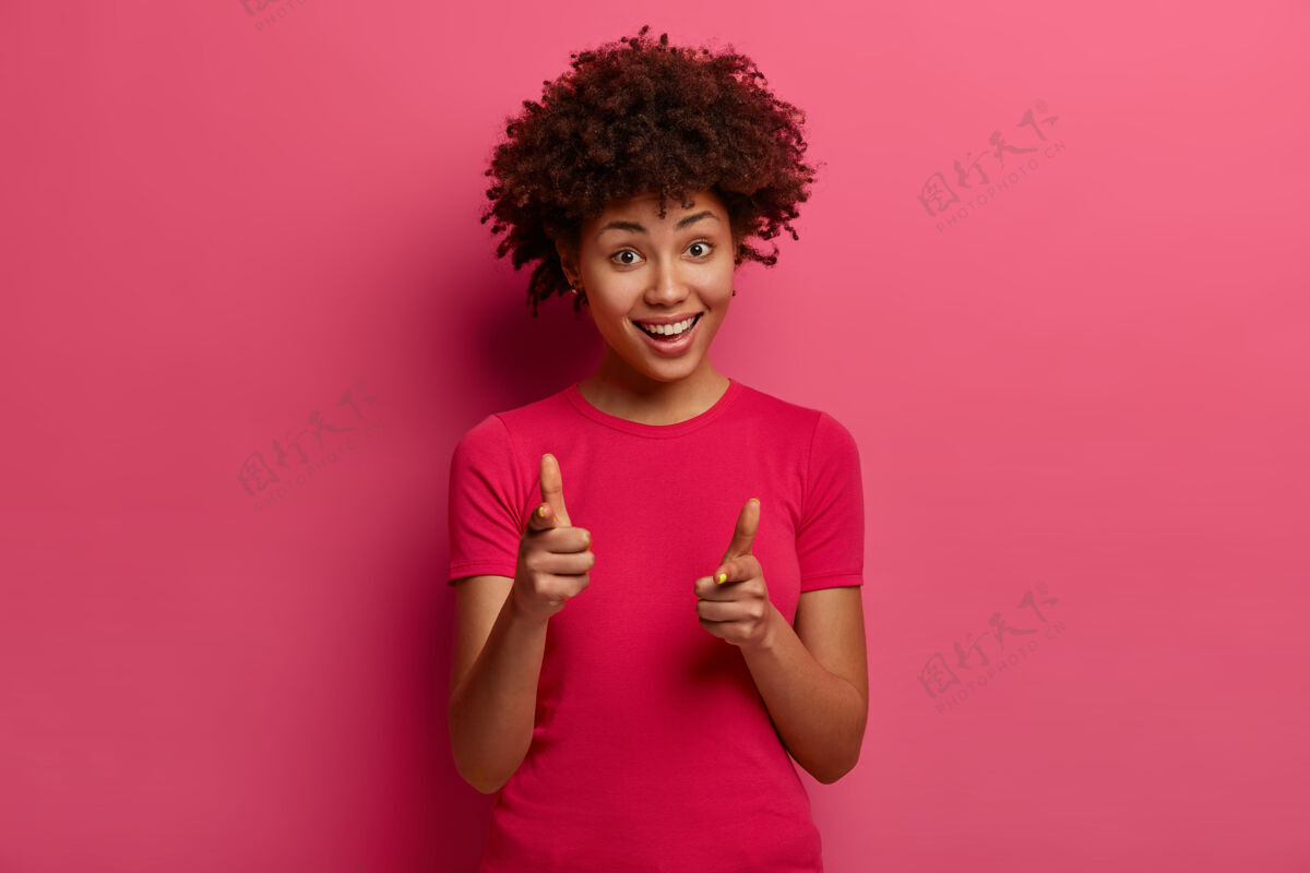 快乐看看吧积极卷曲的女性女孩指着食指说是的 选择了你 开心地微笑 有好玩的心情 穿着随意 对着粉色的墙壁摆姿势满意罗西非洲