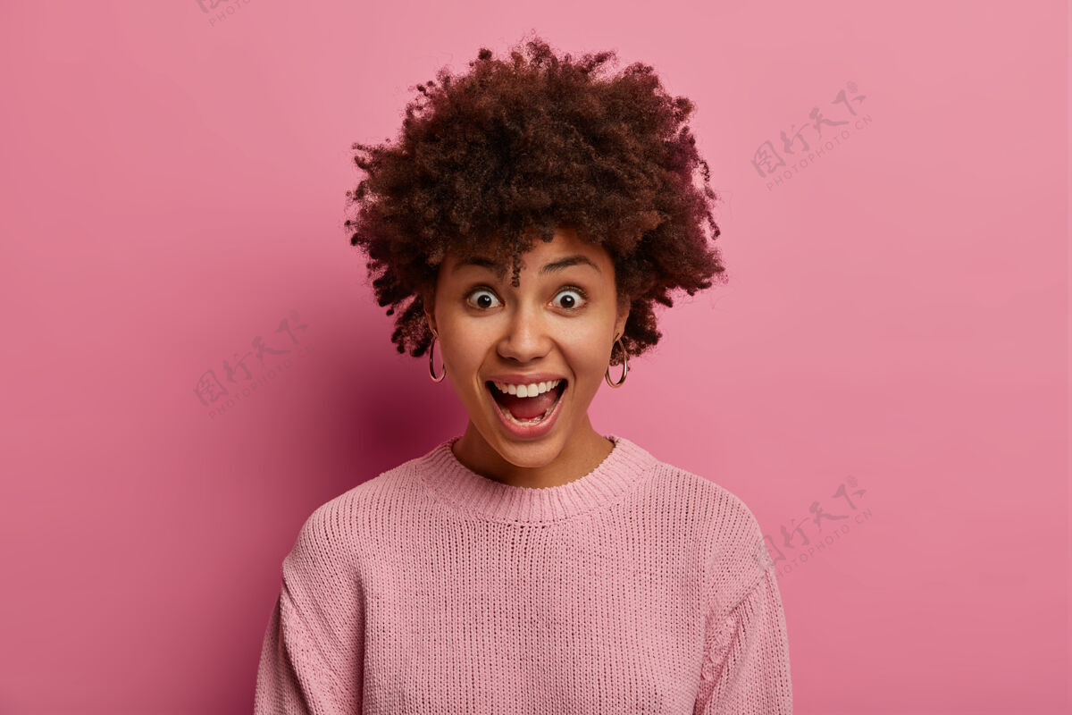 年轻富有感情的非洲发型女人惊呼着 表情惊艳 张大嘴巴 表情欢快有趣 穿着淡粉色的玫瑰色套头衫 在室内摆姿势人啊 兴奋姿势女士天哪