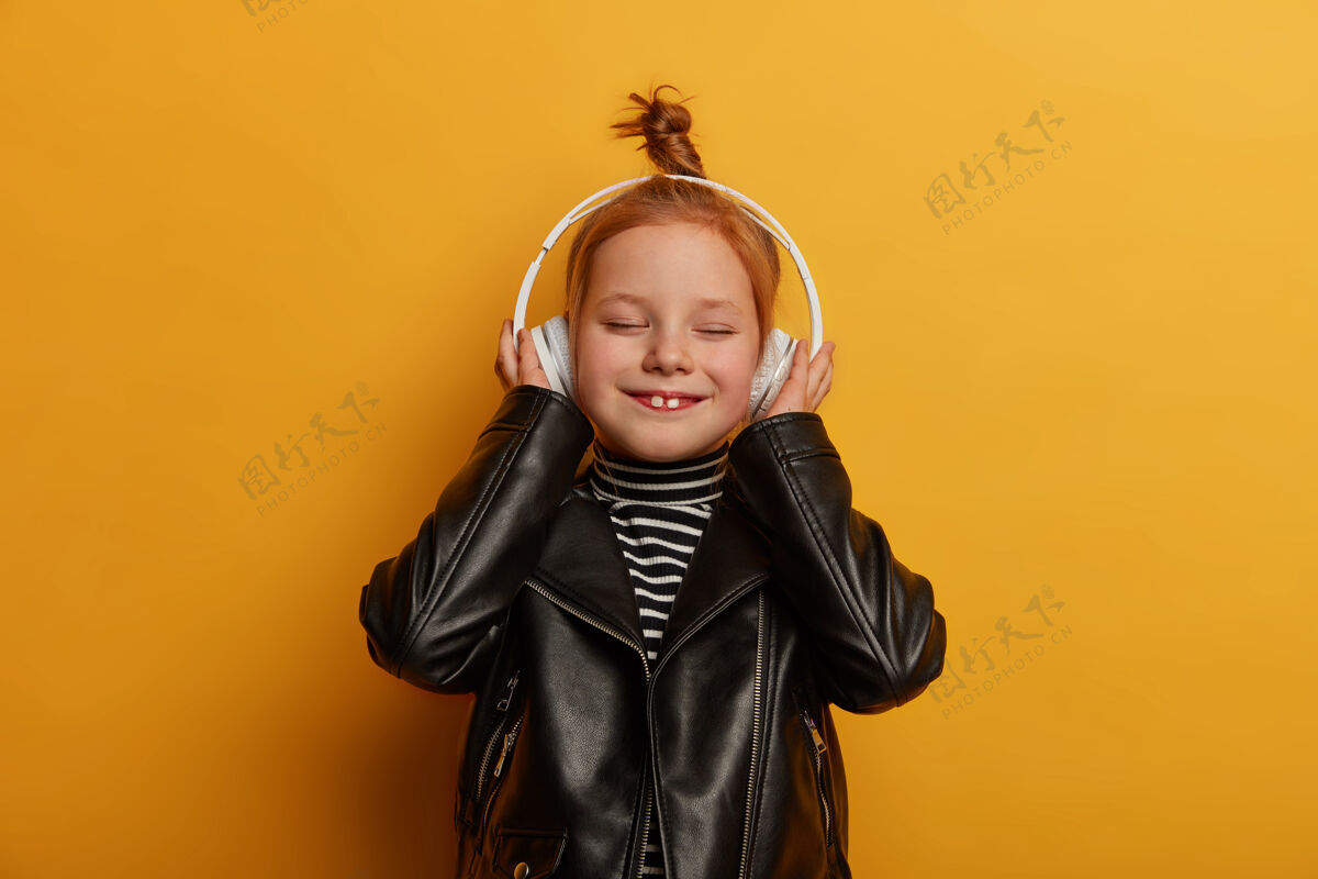 孩子快乐的姜姑娘露出两颗牙齿 戴着耳机听音乐 穿着皮夹克 愉快地闭上眼睛 独自度过空闲时间 隔着黄墙孩子们 娱乐女孩耳机小