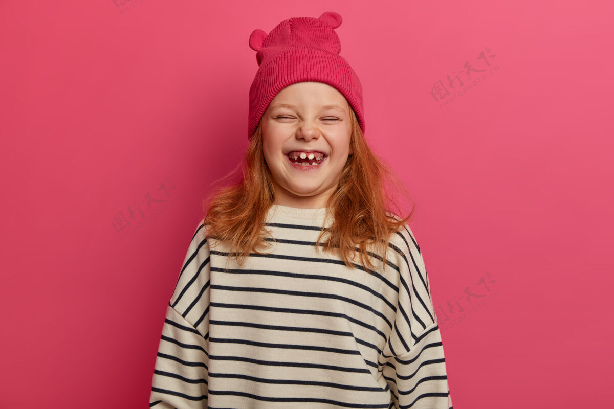积极孩子和快乐的概念快乐的红发女孩从有趣的事情中笑出来 戴着带耳朵的粉红色帽子和宽松的条纹毛衣 笑容灿烂 缺牙 室内模特童年情绪学前班