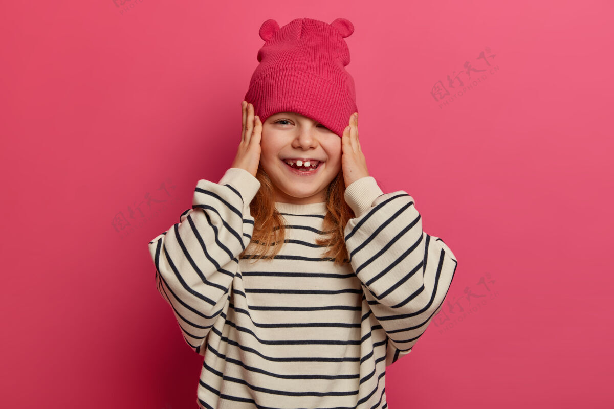 帽子积极进取的女孩很高兴父母给她买了一顶新帽子 穿着宽松的条纹套头衫 满脸笑容 高兴地和其他孩子一起玩 孤立在粉红色的墙上童年 情感人快乐人