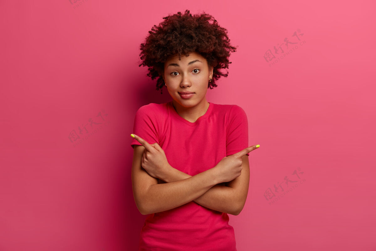 脸被质疑的黑皮肤女人做出选择 显示两个方向 指向侧面 有犹豫的表情 穿着休闲t恤 在玫瑰色的墙上摆姿势 看起来困惑或困惑卷发指标显示