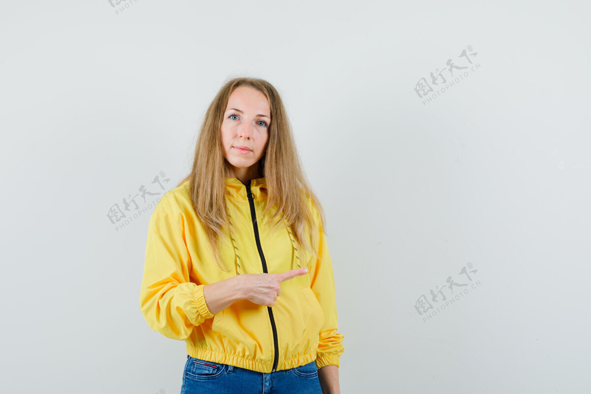 漂亮身穿黄色棒球服和蓝色牛仔裤的年轻女子 指着右边 表情严肃 正对着前方时髦夹克休闲