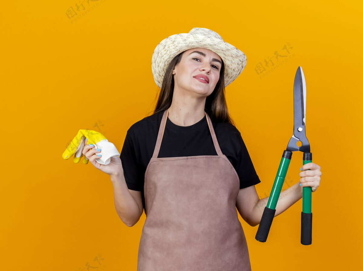 微笑微笑美丽的园丁女孩穿着制服 戴着园艺帽拿着剪刀 手套隔离在橙色背景上花园帽子手握