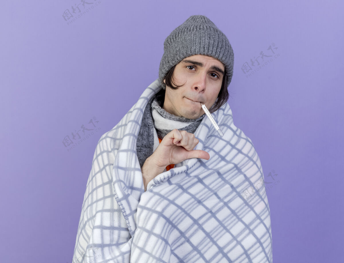 嘴巴未出狱的年轻病夫戴着冬天的帽子 围巾裹在格子布里 嘴里叼着温度计 拇指朝下 孤立在紫色背景上放下帽子年轻人