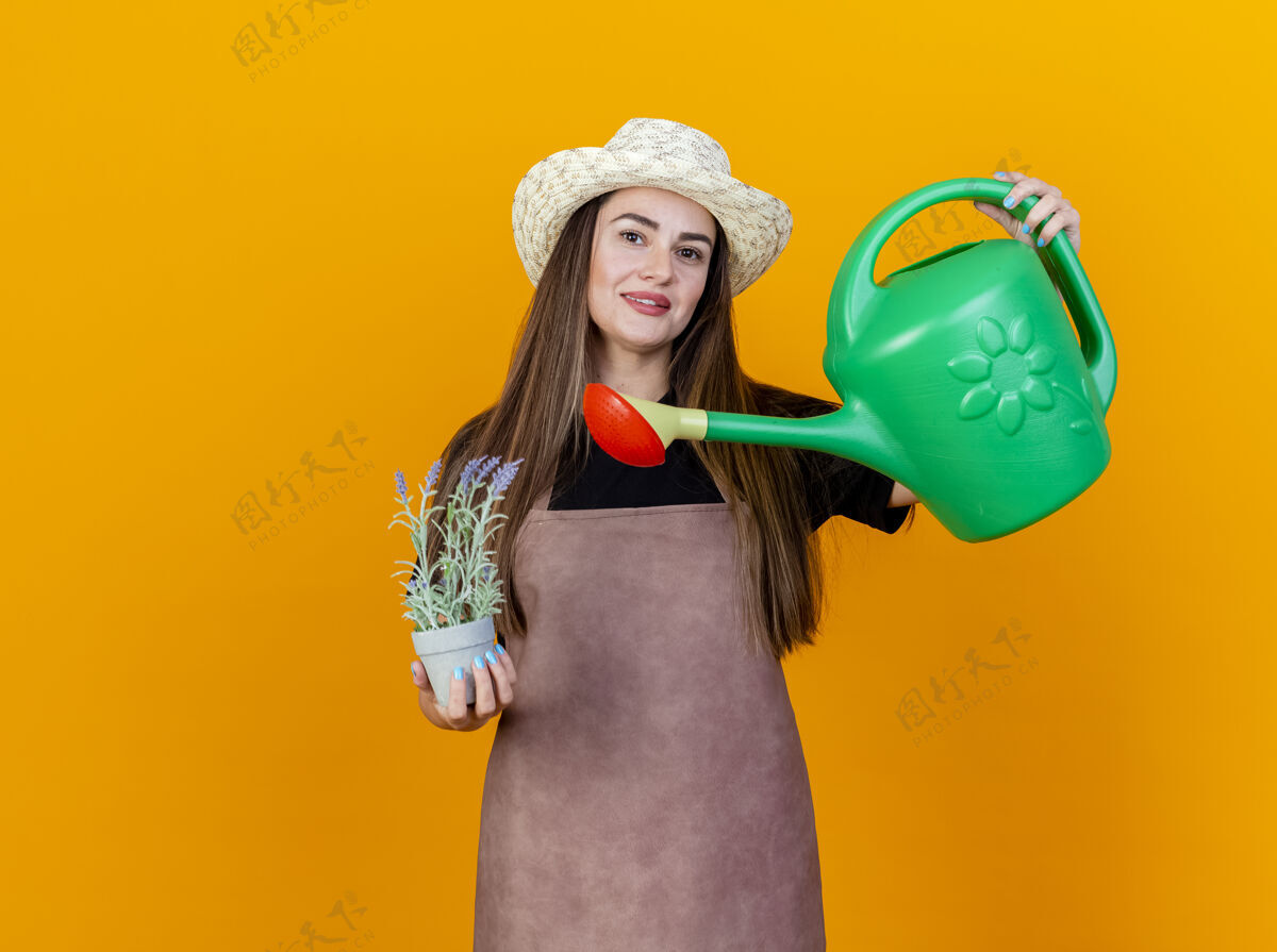 微笑微笑美丽的园丁女孩穿着制服 戴着园艺帽 在花盆里捧着花 在橙色的背景上养着浇水罐浇水制服举行
