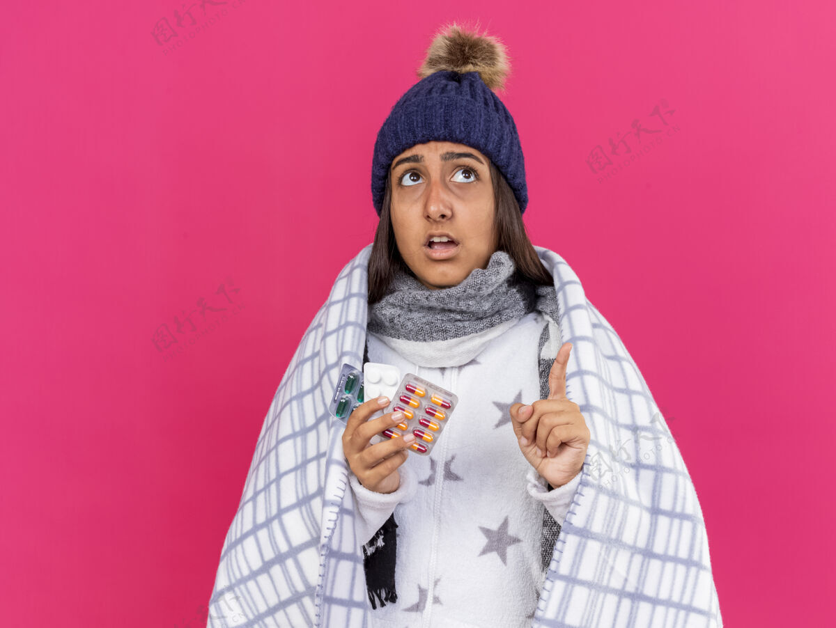 帽子惊讶的年轻生病的女孩戴着冬天的帽子 围巾裹着方格布的药片 指着粉色背景上孤立起来围巾年轻包装
